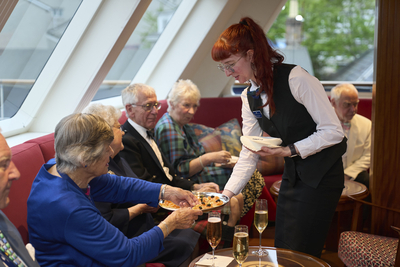Eine Kellnerin serviert Appetithäppchen während eines Aperitifs auf einem Flusskreuzfahrtschiff von Thurgau Reisen. Im Hintergrund sitzen mehrere Passagiere in gemütlicher Runde, genießen ihre Getränke und unterhalten sich. Die elegante Atmosphäre wird durch das stilvolle Interieur des Raumes und die ansprechende Präsentation der Speisen unterstrichen.